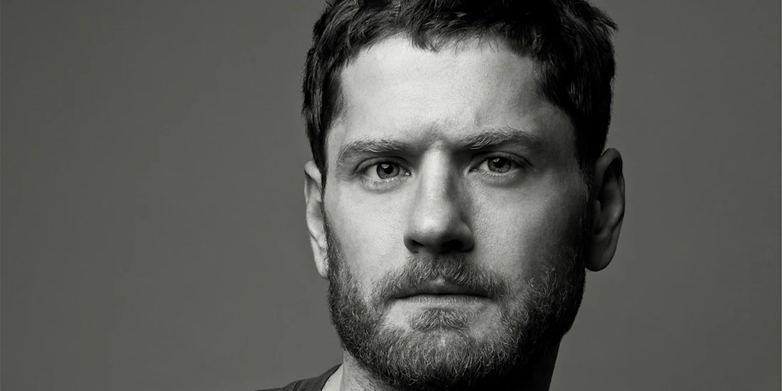 Black and white portrait of a man with short hair and a beard, looking directly at the camera with a neutral expression.