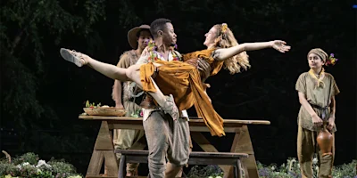 A man in costume lifts a smiling woman in a dress on an outdoor stage, with two other actors in period attire and floral props in the background.