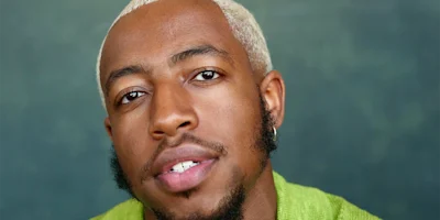 A person with short bleached blond hair, trimmed beard, and an earring looks directly at the camera, wearing a green top against a soft green background.