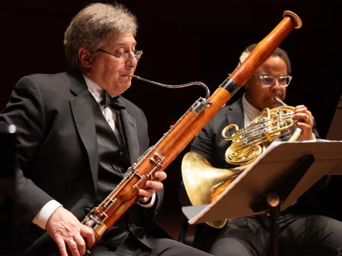 Two musicians in formal attire perform on stage; one plays a bassoon while the other plays a French horn.