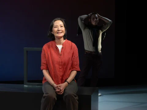 A woman in a red shirt sits smiling on a black bench in the foreground, while a person in a green shirt and scarf stands with hands on head in the background.