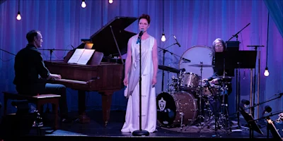 A woman in a white dress sings into a microphone on stage, accompanied by a pianist and a drummer, with stage lights and blue curtains in the background.