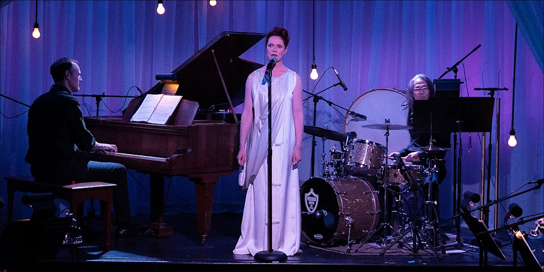 A woman in a white dress sings into a microphone on stage, accompanied by a pianist and a drummer, with stage lights and blue curtains in the background.