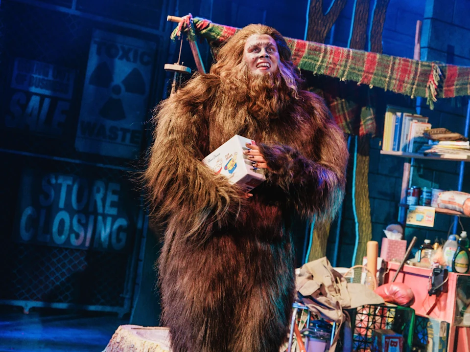 A person in a Bigfoot costume holds a box on a cluttered stage with signs reading "Store Closing" and "Toxic Waste" in the background.