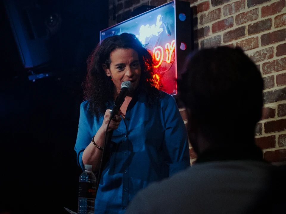 A person with curly hair and wearing a blue shirt speaks into a microphone on stage, with a neon sign and brick wall in the background.