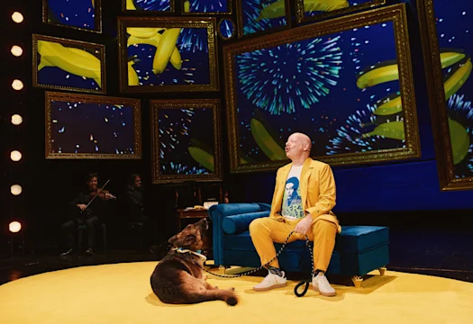 A man in a yellow suit sits on a blue sofa next to a German shepherd, with musicians and digital art featuring bananas and fireworks in gold frames in the background.