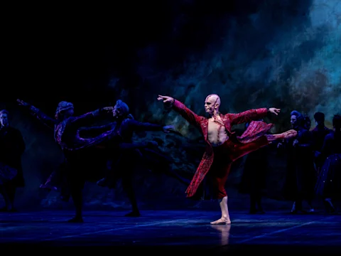 Ballet dancers perform on stage in dramatic lighting, with a central dancer in a red costume extending one leg and arm, surrounded by other dancers in dark outfits.