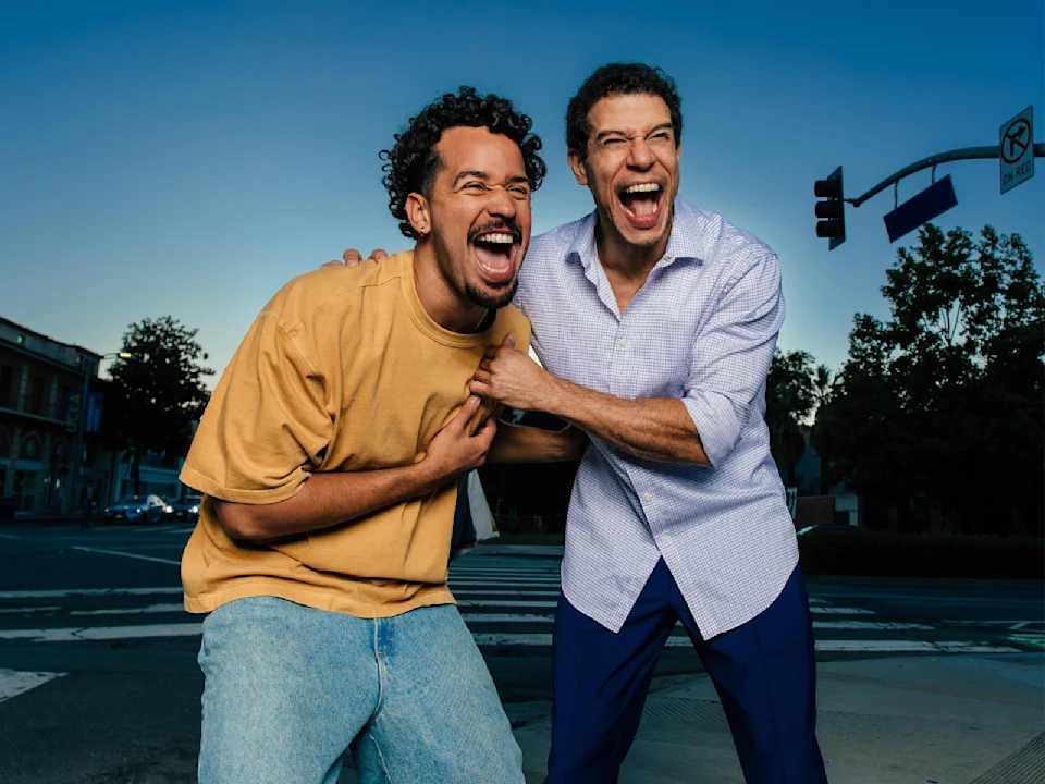 Two men stand on a city street corner, laughing loudly and holding onto each other, with buildings and a street sign in the background.