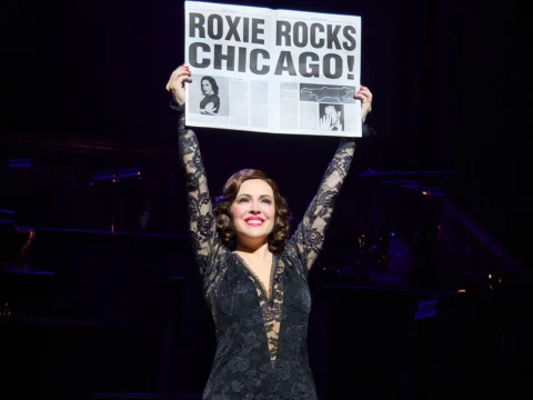 A performer in a black lace outfit holds up a newspaper with the headline "Roxie Rocks Chicago!" on stage.