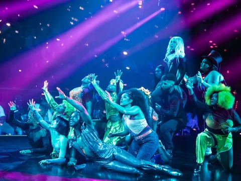 Performers on stage in colorful costumes and dynamic poses under vibrant pink and purple lighting.