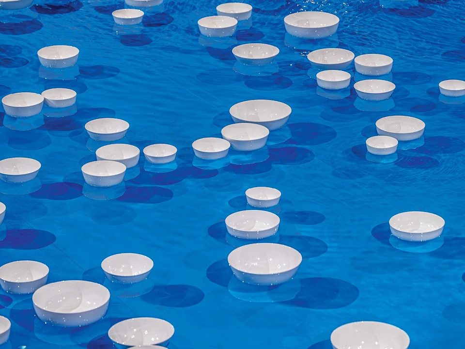 Dozens of white bowls float on bright blue water, casting oval-shaped shadows on the surface.