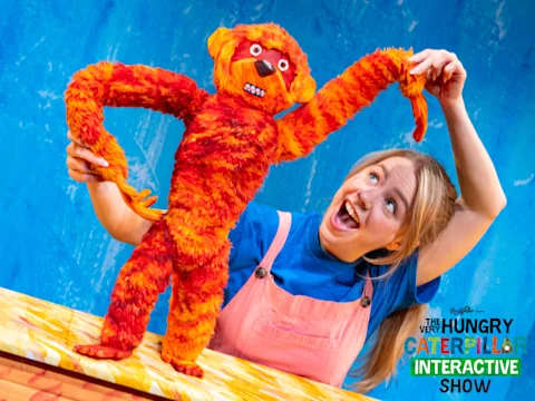 A performer in pink overalls holds up an orange and red animal puppet on a colorful stage with "The Very Hungry Caterpillar Interactive Show" logo in the corner.