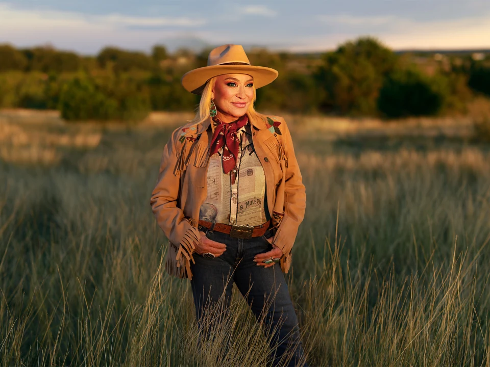 A woman in cowboy attire stands in a field at sunset, wearing a hat, fringed jacket, and jeans, smiling at the camera.