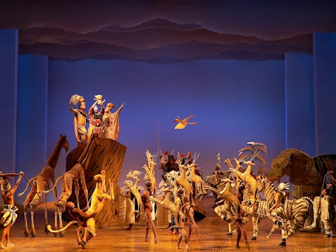 Stage performance featuring actors in elaborate animal costumes and puppets, set against a blue-lit background, portraying a vibrant scene from “The Lion King” musical.