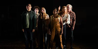 Six people stand in a row facing forward under dramatic lighting, each with a serious or neutral expression, against a dark background.