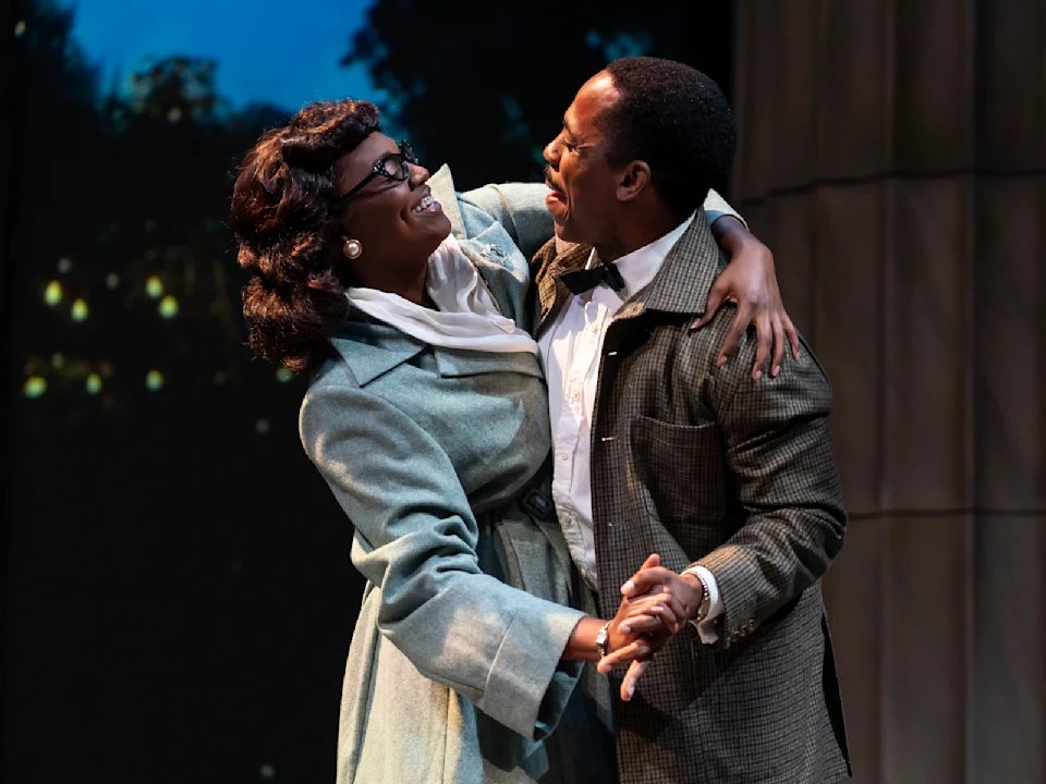 Two people in vintage-style clothing smile and hold each other while dancing, standing in front of a softly lit background with trees.