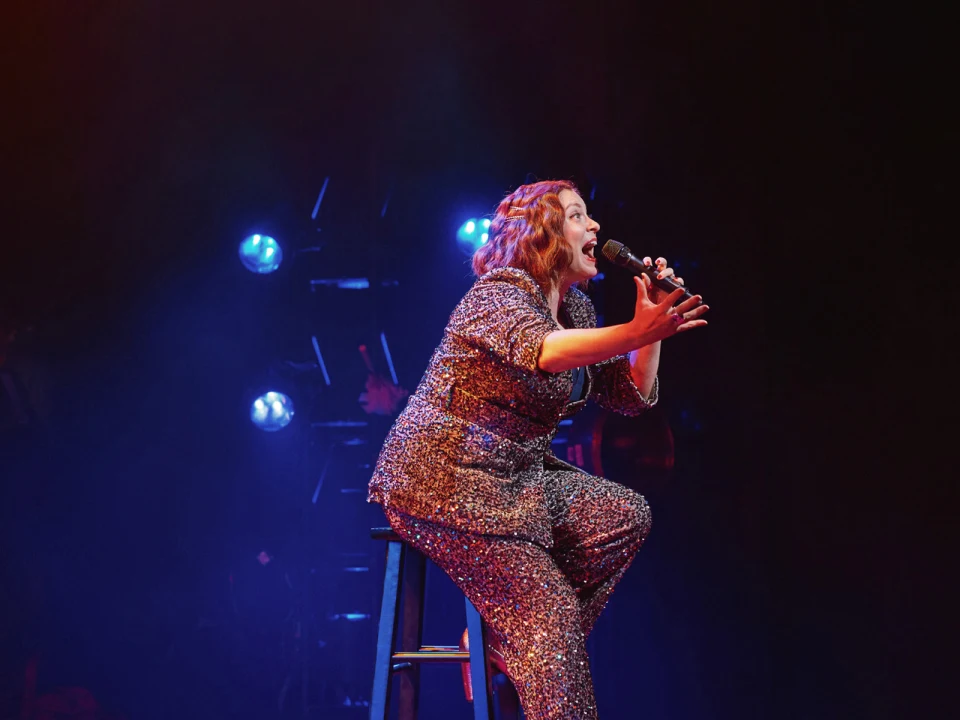 Production of photo of Rachel Bloom: Death, Let Me Do My Show in Chicago with Rachel Bloom in a glittering suit performs on stage, seated on a stool, holding a microphone and gesturing expressively, with stage lights in the background.