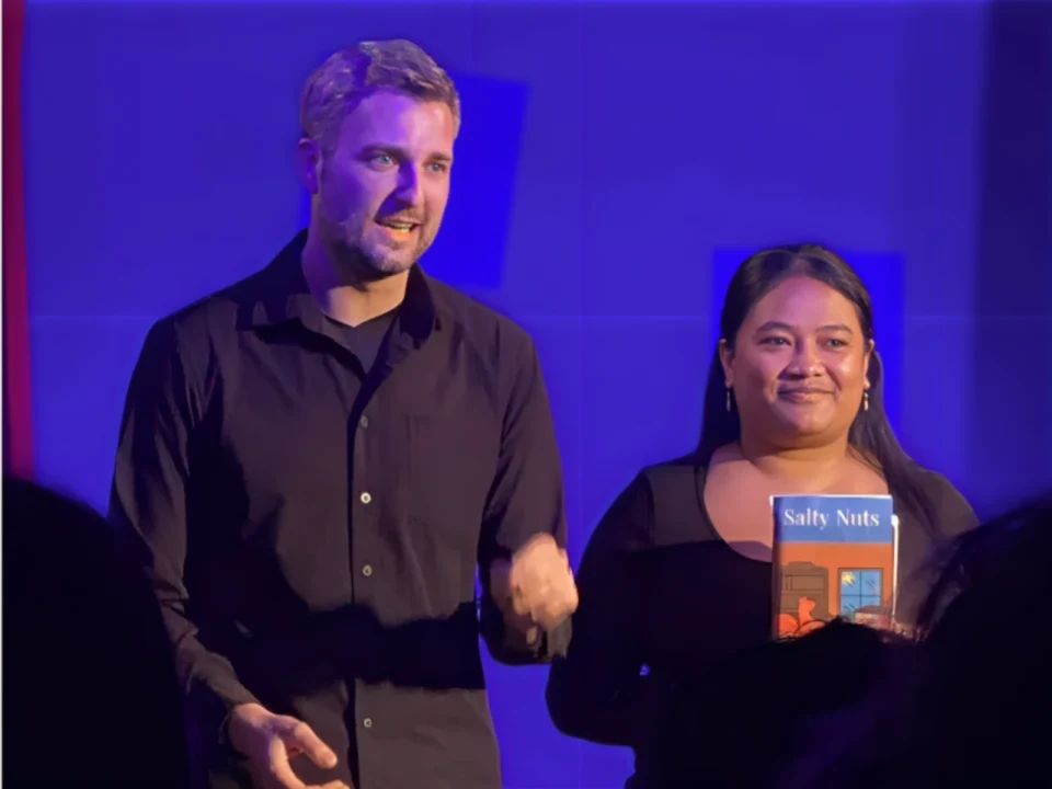 Two individuals stand on a stage, one of them holding a book titled "Sally Notts" and they are both facing the audience.