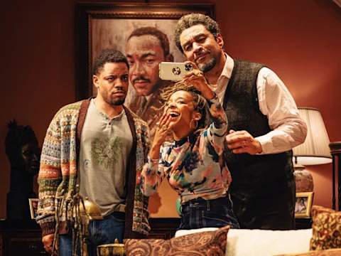 Three people pose for a selfie in a warmly lit living room, with a portrait of a man hanging on the wall behind them. One person looks serious while the others smile.