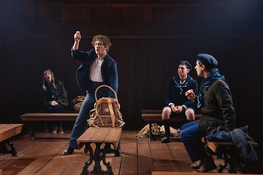 A young man stands and gestures with a raised hand in a wooden room, while three others in blue uniforms sit on benches watching him.