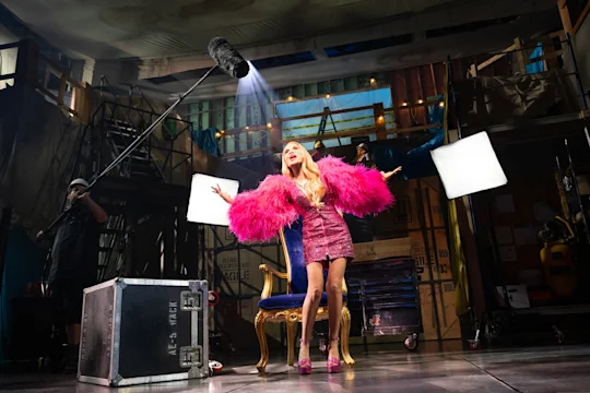 A performer in a pink outfit and feathered sleeves stands on stage in front of a throne-like chair, with studio lights and a boom microphone overhead, during a theatrical production.