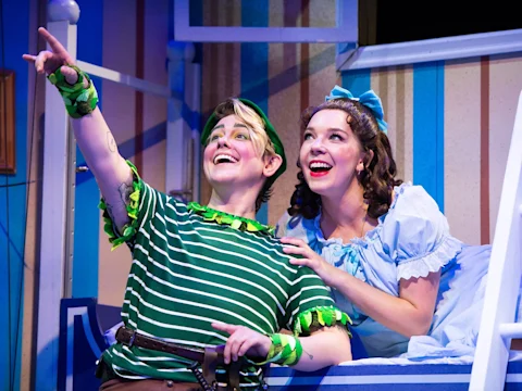 Peter Pan in a striped green shirt points upward as Wendy, in a blue dress, looks on, both smiling during a stage performance.
