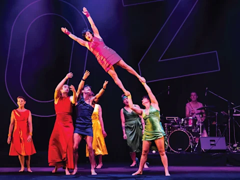 A group of performers in colorful dresses lifts one person above their heads on stage, with a drummer playing in the background.