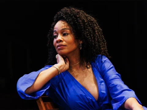 A woman with curly hair wearing a blue dress sits with her hand resting on her neck, looking off to the side.