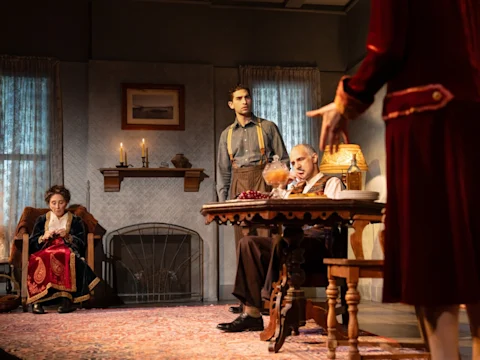 Three people in early 20th-century attire are in a dimly lit living room; one sits by the fireplace, another stands, and a third sits at a table with a woman in red standing nearby.