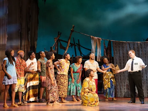 A group of people in colorful attire stand on stage with a man in a white shirt and tie; theatrical set and props are visible in the background.
