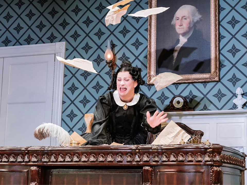 A woman in period costume sits at a desk with papers flying in the air, a quill nearby, and a portrait of George Washington on the wall behind her.