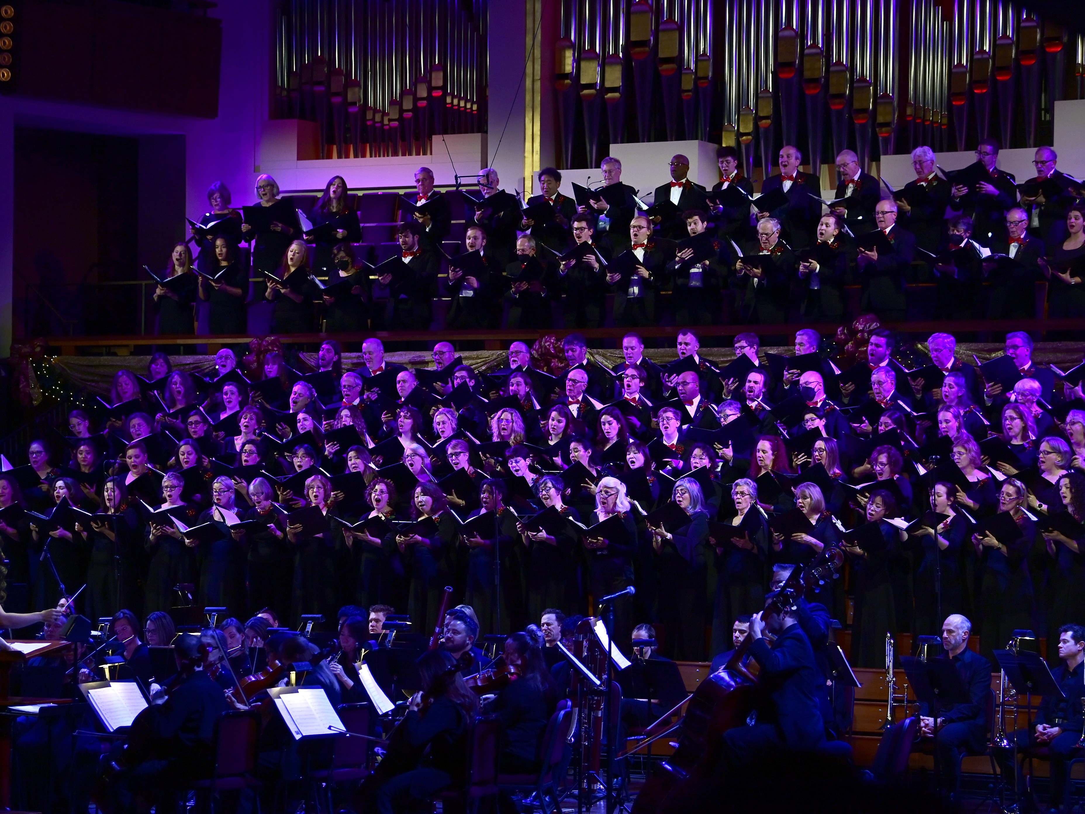 A large choir performs on stage with an orchestra in front of them; an organ is visible in the background under purple stage lighting.