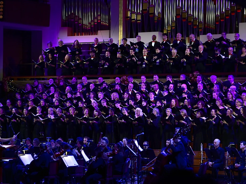 A large choir performs on stage with an orchestra in front of them; an organ is visible in the background under purple stage lighting.