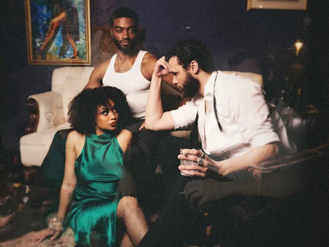Three people are in a dimly lit room; a woman in a green dress sits on the floor, a man in a white tank top stands behind her, and another man in a white shirt sits holding a glass.