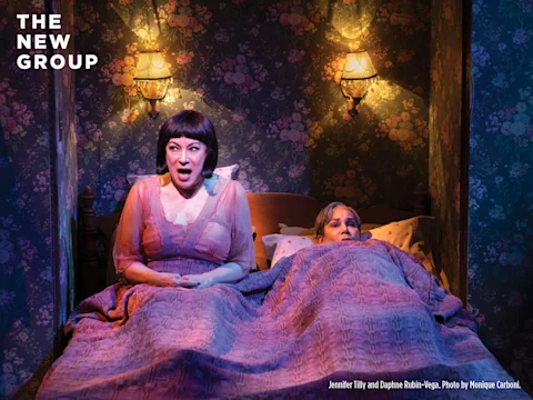 Two women sit in bed under purple blankets in a dimly lit, floral-wallpapered room; one is sitting up speaking, the other is lying down looking concerned.