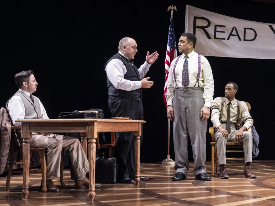 Four men in period clothing are on stage. Three sit, one stands, engaging in conversation under a banner with partial text "READ Y". An American flag is visible in the background.