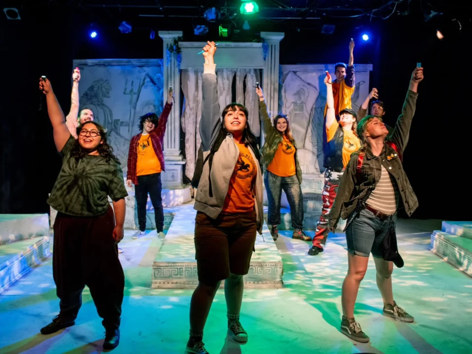 A diverse group of people wearing casual clothing and lanyards stands on stage against a backdrop of white pillars, each with one arm raised, in a theatrical performance with colorful lighting.