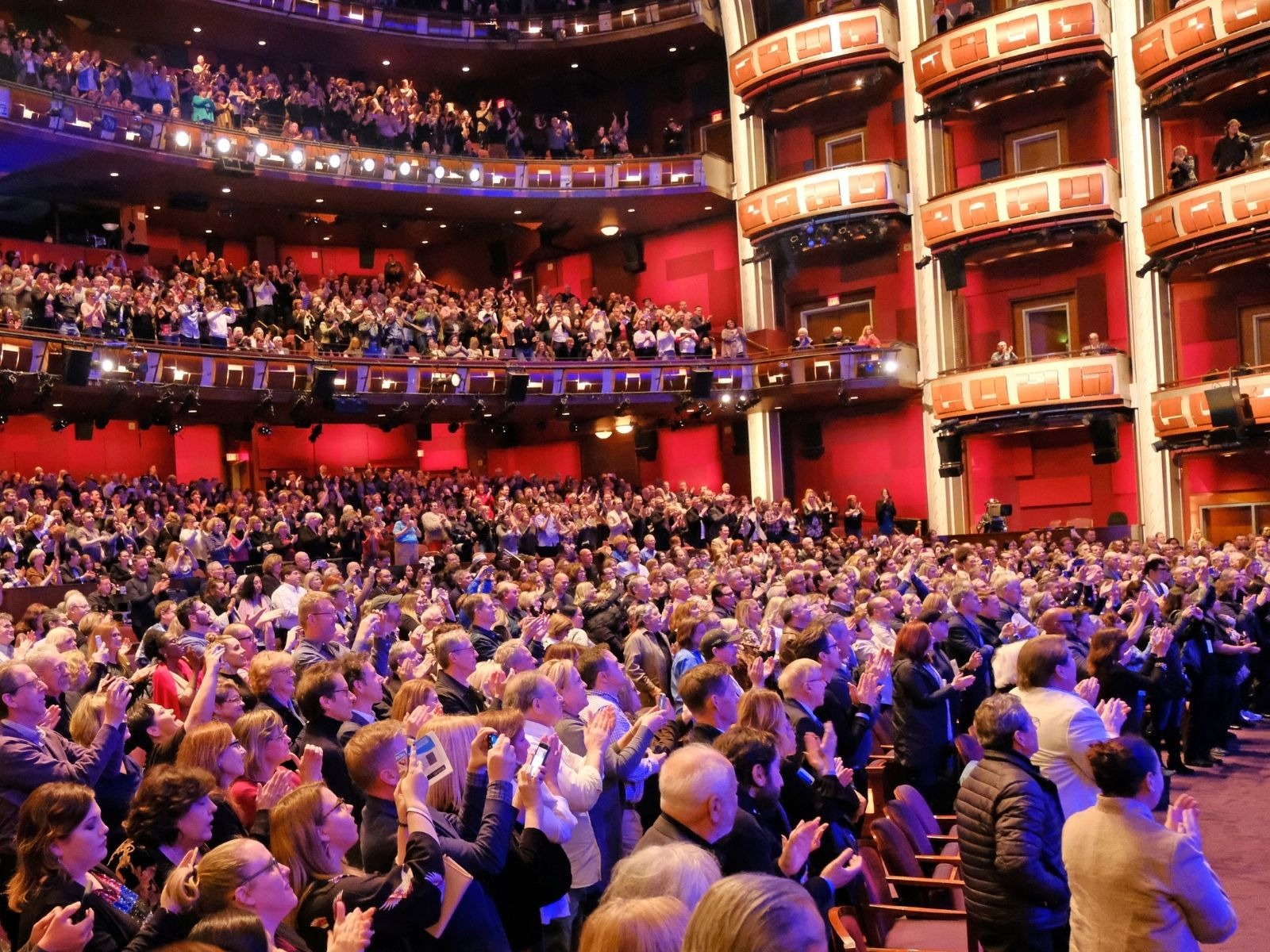 A large audience in a theater stands and applauds, filling multiple levels of seating. The walls are red, and the atmosphere is lively and celebratory.