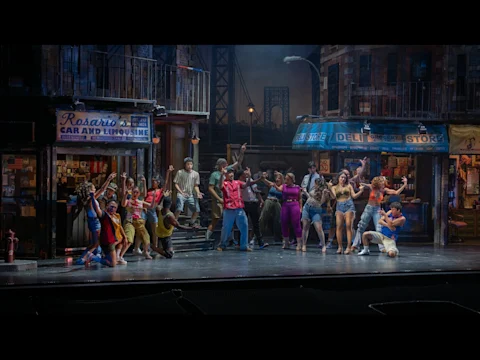 A diverse cast performs a lively group dance on a city street set, with storefronts reading "Rosario's Car and Limousine" and "Deli Grocery Store" in the background.