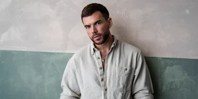 A man with short brown hair and a beard stands against a two-tone wall, wearing a light-colored button-up shirt and looking directly at the camera.