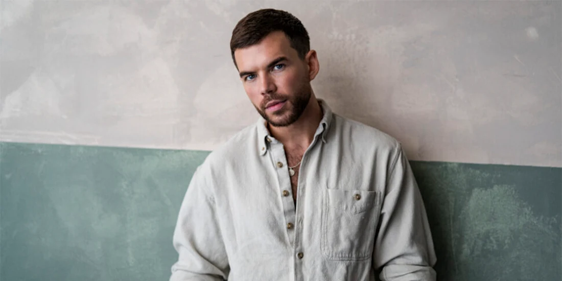 A man with short brown hair and a beard stands against a two-tone wall, wearing a light-colored button-up shirt and looking directly at the camera.