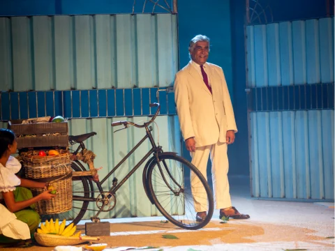 A man in a white suit stands next to an old bicycle with a wicker basket filled with fruits. Another person, partially visible, is seated near the bicycle, handling bananas. The scene is set outdoors.