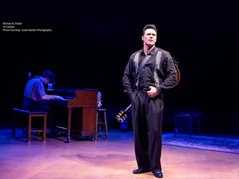 Production photo of Million Dollar Quartet Christmas in Lincolnshire, showing a man in black stands holding a guitar with one hand, looking forward, while another man plays the piano in the background on a dimly lit stage.