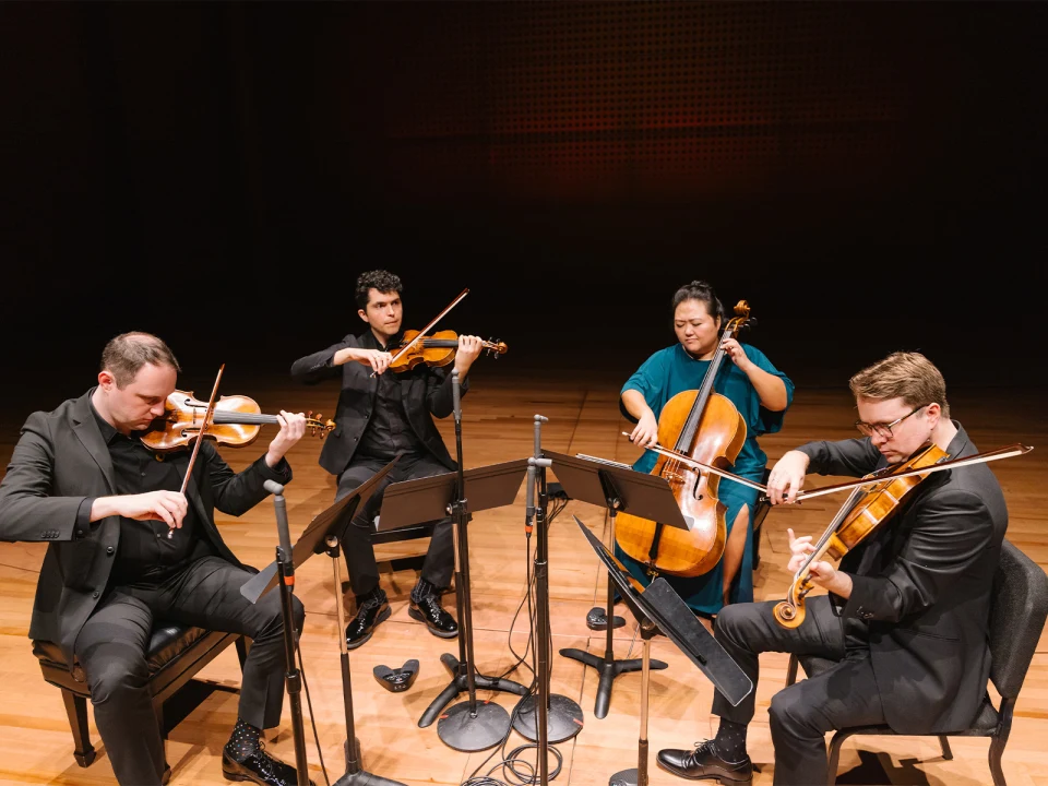 A string quartet performs on a wooden stage. The musicians, dressed in formal attire, play violins, a viola, and a cello, with sheet music stands in front of them.