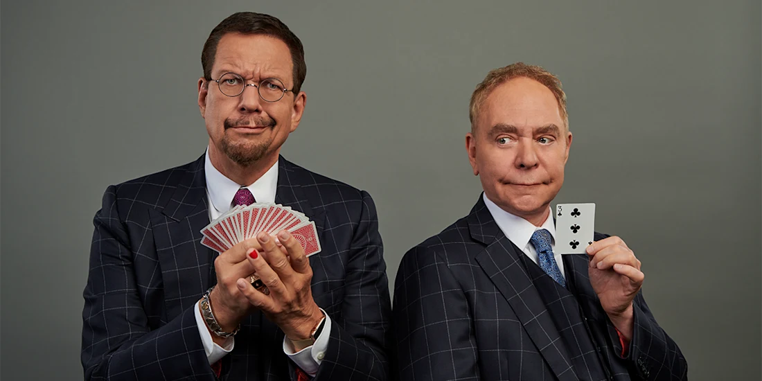 Two men in suits stand side by side; one holds a fan of playing cards, while the other holds up the six of clubs and looks at it with a surprised expression.