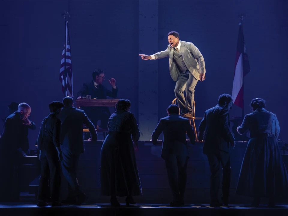 A man in a suit stands on a chair, pointing and speaking passionately to a group of people. Two flags are visible in the background and another man sits at a desk watching the scene.