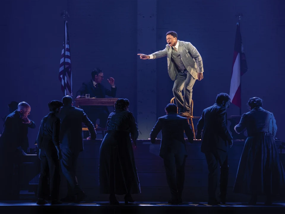 A man in a suit stands on a chair, pointing and speaking passionately to a group of people. Two flags are visible in the background and another man sits at a desk watching the scene.