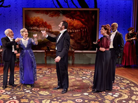 Six actors in period costumes stand on a decorated stage set, raising glasses for a toast; a violinist plays in the background before a scenic mural of trees.