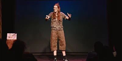A person wearing a leopard print outfit stands on stage holding a microphone, speaking animatedly to an audience.