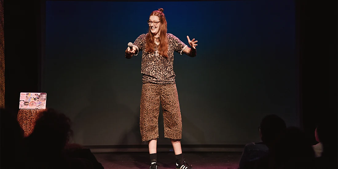 A person wearing a leopard print outfit stands on stage holding a microphone, speaking animatedly to an audience.